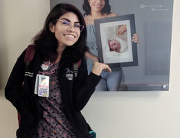 Nurse Laura stands in front of hallway image of herself holding a photo of newborn Laura when she was in NICU