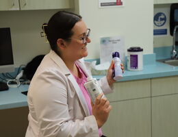 A woman is holding up two protein shakes as she talks to a patient in a medical room.