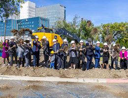 Hospital admin wearing hard hats stand in line in front of contruction vehicle and shoveling the ground