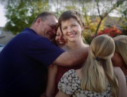 family surrounds teenage daughter, all in a group hug outside smiling broadly
