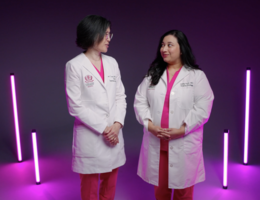 Two women stand shoulder to shoulder with pink light fixtures standing behind them. Both women are wearing pink scrubs with white doctors coats. Woman on left is Asian and woman on right is middle-eastern. 