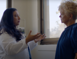 Doctor with brown hair in white coat has a conversation with a patient wearing blue top with short blonde hair. 