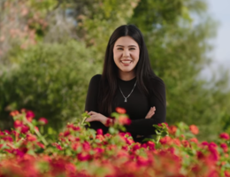 Young woman with dark hair and a black shirt stands smiling with her arms crossed in front. Red flowered bushes are in the foreground. 