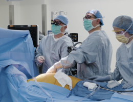 Three people in blue scrubs stand over operating table looking to the viewers left.