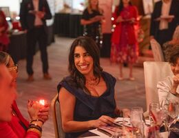 Guests sit at an elegantly set table during a gala event, surrounded by warm lighting, glassware, and other attendees in the background.