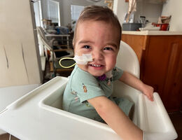 one year old boy sitting in high chair, smiling at camera, medical tube in nose