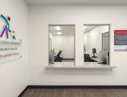 Reception area at Loma Linda University Children’s Health Pediatric and Adult Medicine clinic with two service windows, a counter, office workstations visible behind glass, and clinic signage on a bright, modern interior wall.