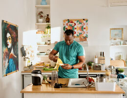 An African-American male in a bright kitchen prepares a banana at a wooden countertop surrounded by cooking tools, fresh produce, and a blender, with colorful artwork and neatly arranged shelves in the background.