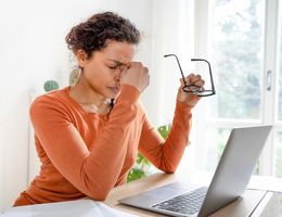 Woman sitting, pinching top of her nose holding eye glasses in front of a laptop