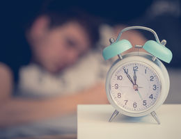 Alarm clock on nightstand in foreground, person sleeping in the background