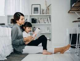 Young Asian mother sitting on the floor in the bedroom reading book to little daughter, enjoying family bonding time together at home