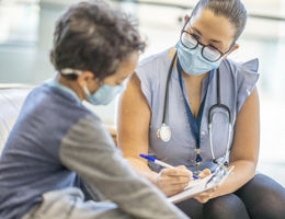 A Hispanic female doctor is taking down notes while speaking to a young 8 year old male patient. Both doctor and patient are wearing masks during the coronavirus pandemic.