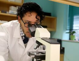 A doctor in a white lab coat and black hair looks through a microscope