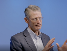 a man in front of a blue background is talking with his hands up 