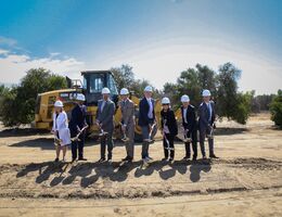 eight hospital leadership representatives dig dirt during the groundbreaking ceremony in front of a dump truck with trees in the background.