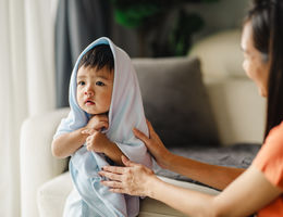 Asian boy with a towel on his head is sitting in the living room. He has big, dark black eyes and is relaxing on the sofa after his bath.