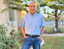 Man in blue button up and jeans stands in front of his house with his hands in his pocket