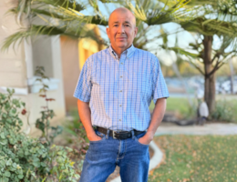 Man in blue button up and jeans stands in front of his house with his hands in his pocket