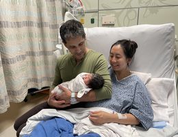 Married couple sits on bed together holding their newborn baby